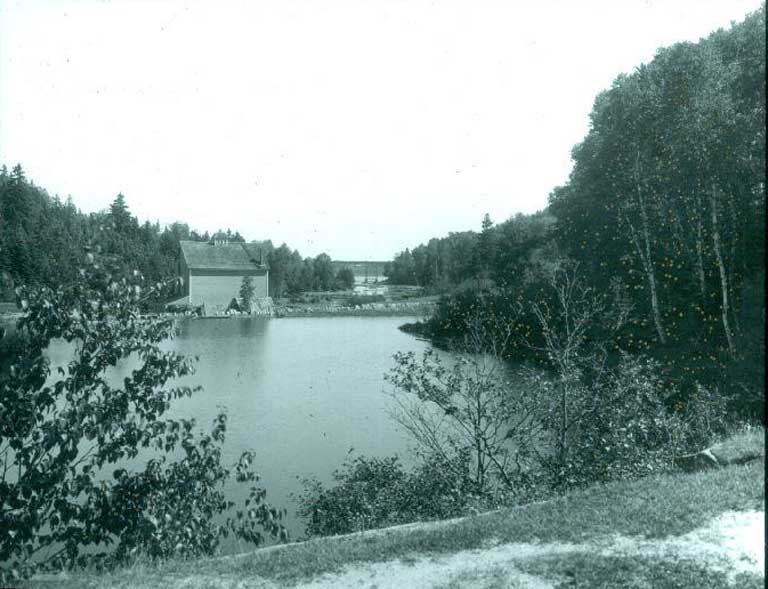 Photographie noir et blanc d’un lac et d’arbres avec un ancien bâtiment en bois