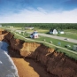 Colour photograph of a shoreline with brown cliffs and green fields on top with a lighthouse building
